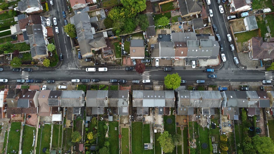 An aerial view of a residential street with tightly packed terraced houses, parked cars lining both sides of the road, and small back gardens visible behind each property. The image captures a busy ur