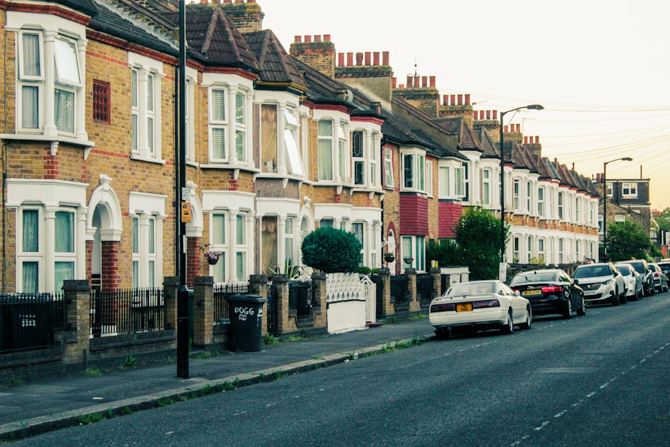 An aerial view of a residential street with tightly packed terraced houses, parked cars lining both sides of the road, and small back gardens visible behind each property. The image captures a busy ur